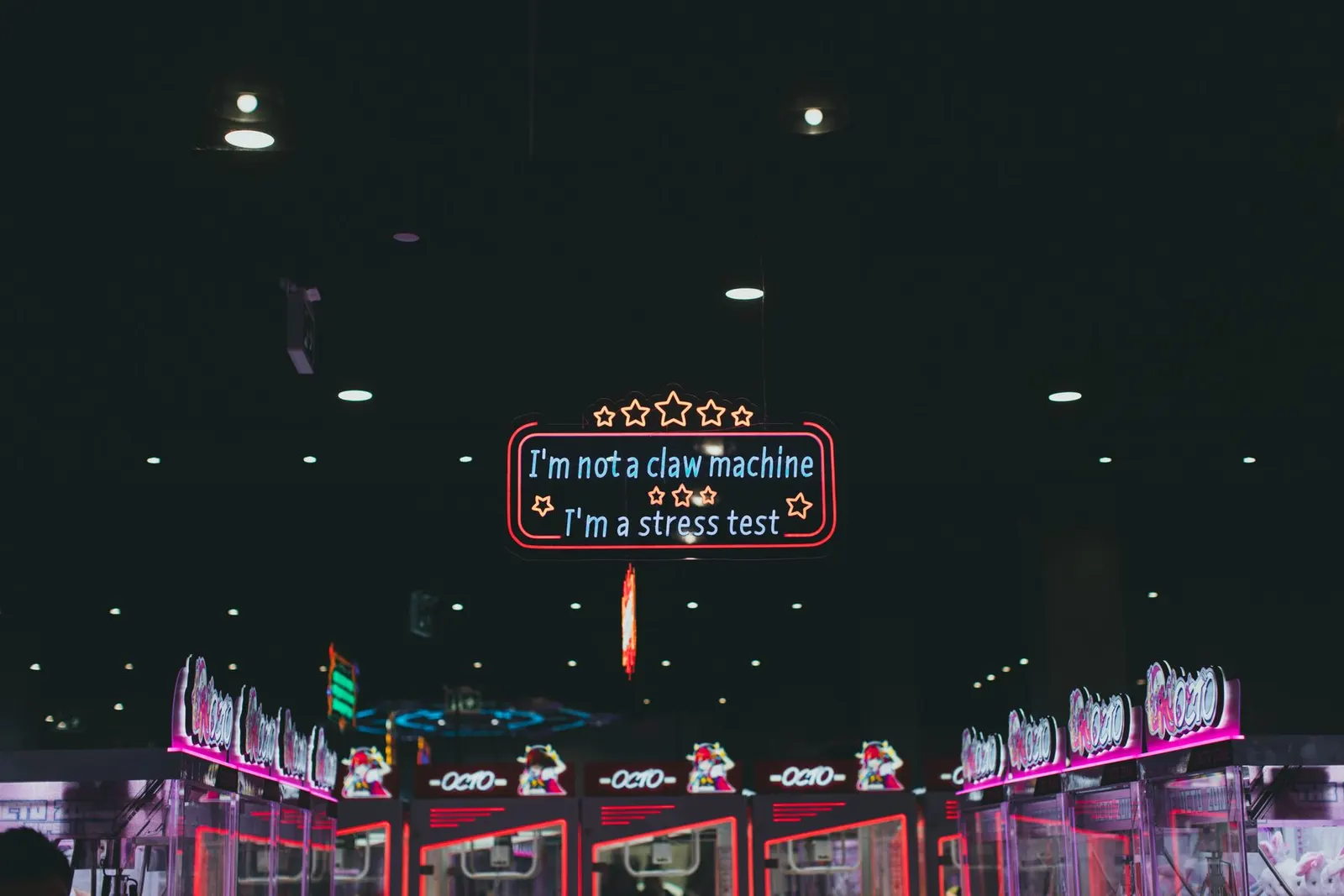 A dark arcade lit by pink and red neon, with a glowing sign reading I'm not a claw machine, I'm a stress test