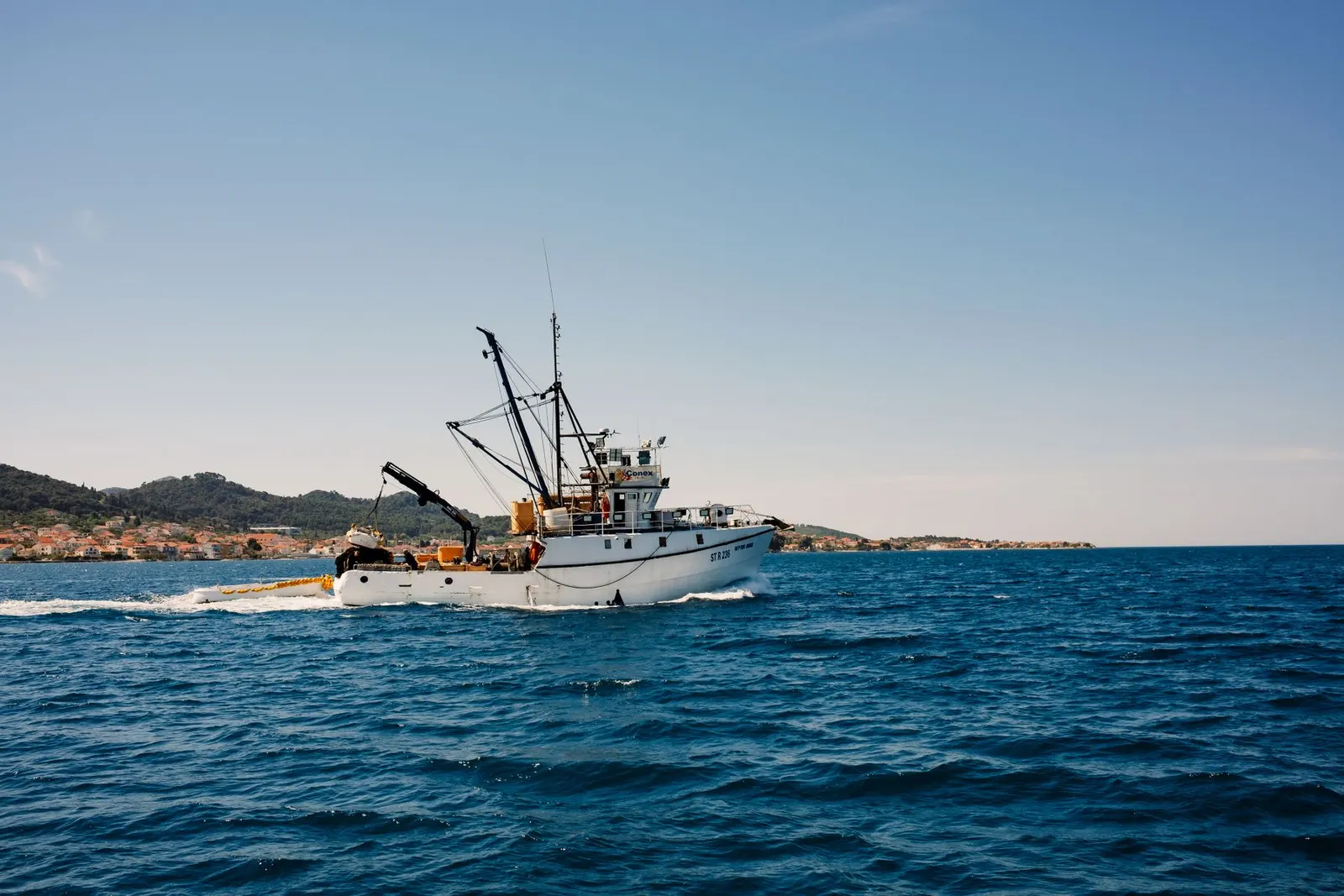 A fishing trawler sails across the open sea with distant hills and a town on the horizon